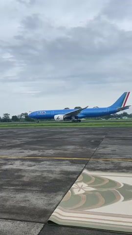 A video shows a blue ITA Airways aircraft taxiing on a paved runway, viewed from a slightly elevated position. The runway is marked with yellow lines and shows signs of wear. The sky is overcast, with grey clouds filling the frame. In the foreground, a rolled-up mat with a floral pattern is visible on the ground. Trees and greenery are visible in the background, beyond the runway. A sound of marching band music is audible.