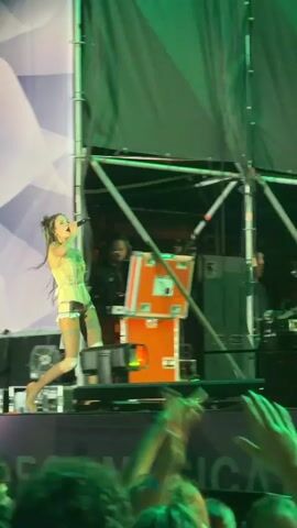 This video captures a live music performance on an outdoor stage, likely in Segrate, Italy, given the context. The main subject is a female singer in the foreground, dressed in a distinctive outfit: a light-colored, structured corset or bustier paired with what appear to be high-waisted, brightly colored (possibly orange and white striped) pants or shorts, and knee-high legwear. She has long, dark hair and is actively engaging with the audience, holding a microphone and gesturing with her hands. Her expression suggests she is performing with energy and enthusiasm.

Behind the singer, there are other individuals, including a person behind a keyboard or DJ setup, and others less clearly visible in the background. The stage is constructed with scaffolding and draped with a large green fabric that serves as a backdrop. Various technical equipment, including speakers and lighting rigs, are visible. Red and orange stage lights are illuminating the performer, creating a dynamic visual effect against the green backdrop. The overall atmosphere appears to be a lively concert or festival setting, with the audience visible in the foreground, some holding up phones to record or photograph the performance. The presence of these recording devices and the raised hands indicate audience engagement. The time of day is likely evening or night, as indicated by the artificial stage lighting. Visible text on the stage edge, partially obscured, includes the word "MUSICA".