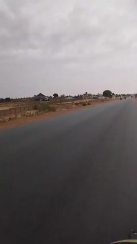 The video shows a black asphalt road stretching into the distance under a cloudy sky. The land adjacent to the road is sandy and sparsely vegetated, dotted with trees and low-rise buildings. As the video progresses, two motorcycles, each carrying two people, approach and pass the camera. The environment appears arid and the scene suggests a rural or semi-rural setting.