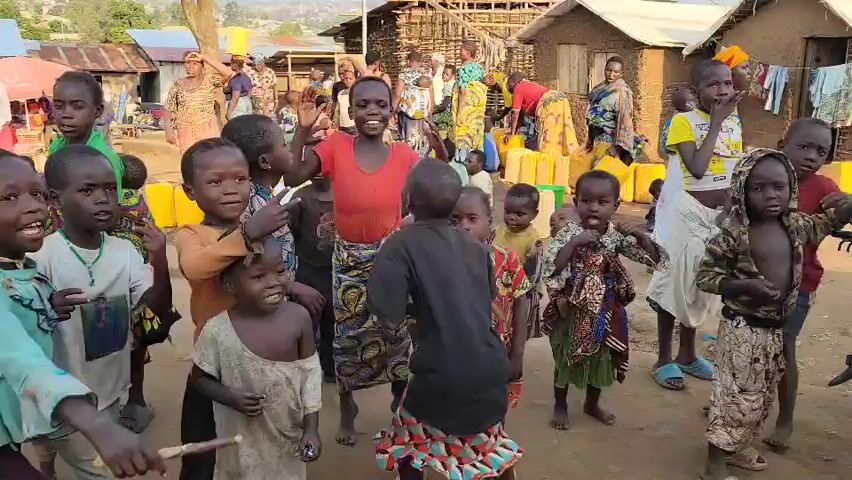 A group of children are gathered in a dusty outdoor area with several adults in the background. The children are varied in age and are dressed in colorful clothing. Some of the children are smiling and looking at the camera, while others are interacting with each other. In the background, there are small buildings that appear to be made of wood and mud. There are also yellow containers visible, possibly for water. Two adults are positioned in the foreground, facing away from the camera, with one of them appearing to be interacting with the children. The overall scene suggests a community setting, possibly in a developing country.