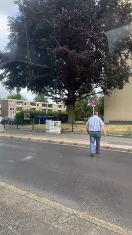 A man in a blue and white plaid shirt and blue jeans walks away from the camera on a sidewalk. Behind him is a tree with dark foliage, apartment buildings, and a street with cars. The weather appears to be overcast.