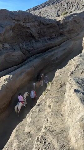 The video shows a group of people riding horses down a steep, narrow dirt path between rocky cliffs. The path is a ravine or canyon with rough, uneven terrain. The people are dressed in casual clothing. The horses are moving slowly down the path, kicking up dust. The sky is visible at the top of the frame and appears to be a clear blue. The scene suggests an outdoor activity in a natural, rugged landscape.