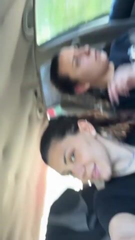 Two women are in a car, presumably as passengers. The camera angle is from below, pointing up at them. The woman closest to the camera is singing along to music playing in the car and holding the phone. She is wearing a black t-shirt, a blue beaded necklace, and has her hair pulled back. Her face has freckles and she appears to be wearing light makeup. The other woman is in the background, looking up at the car's ceiling. She is also wearing a black t-shirt and has her hair slicked back. The car is moving, with trees visible through the window.