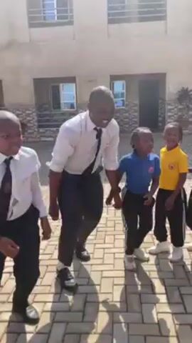A video shows a group of five children and an adult male dancing outside in a schoolyard. The building behind them is two stories tall, light-colored, and has many windows. The ground is paved with tan bricks. The adult male and two of the children are wearing school uniforms of white shirts, black ties, and black pants. The other three children are wearing athletic uniforms in bright colors. The adult male is in the middle of the group, smiling and leading the dancing. The children are smiling and appear to be enjoying themselves. The lighting is bright and sunny.