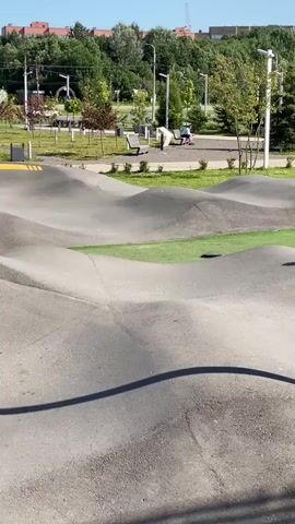 This video captures a bright, sunny day at a skatepark designed for scooters and bikes, likely located in Cherepovets, Russia. The park features a series of undulating concrete ramps and curves, ideal for pump track riding.

The primary subjects are people engaged in activities at the park. In the foreground, a young person on a scooter is actively riding on the track. Throughout the video, other individuals, including children and adults, can be seen in the background, some spectating, others riding bikes or scooters, and some relaxing on park benches. The setting is an outdoor public park with green spaces, trees, and modern urban apartment buildings visible in the distance. The time of day is clearly daytime, with bright sunlight casting distinct shadows, suggesting it's mid-morning to afternoon. The weather appears to be clear and warm.

Notable details include the distinct yellow markings on the concrete ramps, which likely serve as directional cues or visual aids for riders. The overall mood is one of recreation and enjoyment, with people participating in sporting activities in a well-maintained outdoor environment. No visible text is discernible in the video.