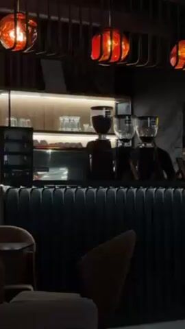 The camera slowly pans right, revealing a coffee shop. Dark, cylindrical lights hang over a black counter. Three coffee grinders sit on the counter, to the left of a cash register and computer monitor. Behind the counter, a shelf holds glasses. To the right, a neon sign reads "something sweet." Seating is visible in the foreground, with brown chairs and tables.