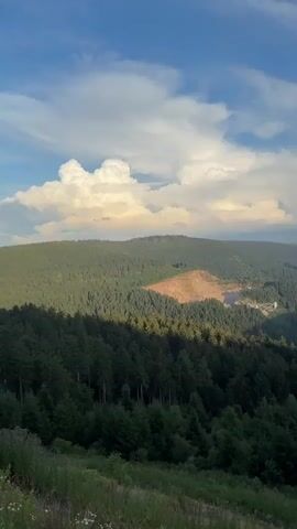 This is a vertical, handheld video of a mountainous landscape covered in dense, green forests under a partly cloudy blue sky. The camera pans slowly from left to right. In the foreground, there is a grassy slope with some dry vegetation and a faint shadow. In the mid-ground, the vast expanse of evergreen trees dominates the rolling hills. Towards the center-left of the frame, a large, lighter-colored patch of earth indicates a cleared area or construction site, with some buildings and a body of water visible within it. The background shows layers of distant, hazy mountains fading into the horizon. The lighting suggests it is late afternoon or early evening, with sunlight casting shadows and highlighting some of the clouds with a warm glow.