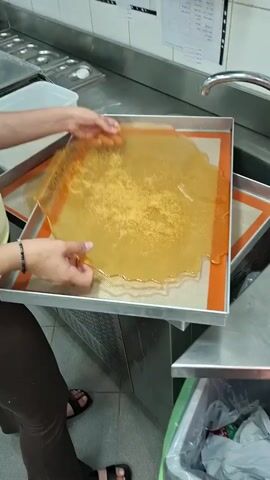 Here's a description of the video: 

A person is shown breaking apart a large, thin sheet of brittle amber-colored candy. The candy is being broken over a rectangular metal baking tray lined with a silicone mat. The person is wearing dark pants and sandals and a bracelet. The scene appears to be in a commercial kitchen, with stainless steel surfaces and a sink visible in the background. There's also a wastebasket nearby. The brittle breaks easily, creating smaller pieces on the tray.
