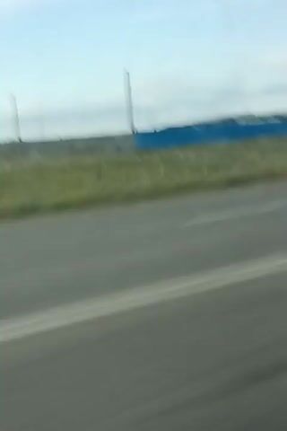 The video shows a blurry, fast-moving shot of a road with a few buildings and a blue fence in the background. The sky is visible, appearing overcast or hazy. The camera appears to be inside a moving vehicle, likely a car, looking out a window. The buildings are industrial or commercial in appearance, with one appearing to be a light-colored, two-story structure. There are no people visible in the footage. The environment appears to be rural or semi-rural, with some grass and unpaved ground visible.