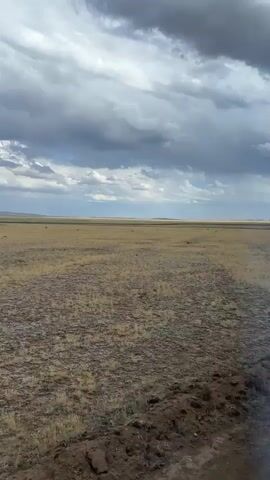 This media file captures a view from inside a vehicle, likely a bus, looking out at a vast, open landscape. The scene is dominated by a wide, flat plain covered in dry, yellowish-brown grass and sparse vegetation. The horizon is distant and flat, with faint outlines of hills or mountains in the far distance.

The sky above is dramatic, filled with heavy, dark grey clouds that suggest impending rain or a storm. Patches of lighter sky are visible between the clouds, indicating it's daytime, likely late afternoon or early evening given the lighting and cloud cover. The overall mood is somewhat bleak and desolate due to the overcast sky and the arid landscape.

The foreground and right side of the frame show elements of the vehicle's interior. A dark fabric, possibly a seat or a bag, is prominently visible in the lower right and center. A bright blue curtain with a subtle patterned texture is drawn back along the right side, revealing the window and the exterior view. Above the curtain, a section of the vehicle's ceiling is visible, showing a row of overhead storage compartments or lights, with some blue plastic elements and metal fixtures. There is no visible text in the media file. The location context of Bakty, Kazakhstan, suggests a remote, possibly steppe-like environment.