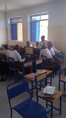 The video shows a classroom setting, likely a computer lab, where a group of students are working on computers. The students are seated at individual desks, each equipped with a desktop computer. The computers are on, displaying content. The students are dressed in school uniforms, consisting of white shirts and brown skirts or trousers. The room has two windows that provide natural light. A teacher is standing, observing the students. There are also stacks of books on some of the desks. The overall impression is of a typical classroom environment where students are engaged in a computer-based learning activity.