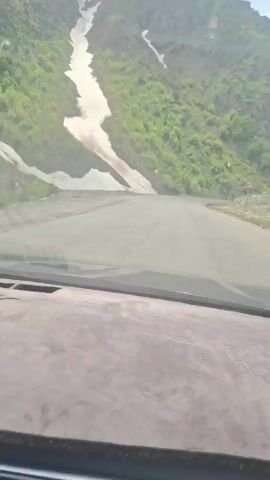 A first-person perspective video shows a car driving on a paved road through a mountain pass. The mountain to the left has a significant patch of snow on its side, surrounded by green vegetation. The sky is partly cloudy. A silver car drives past in the opposite direction. There is a road sign on the right side of the road. The dashboard of the vehicle recording the video is visible in the foreground.