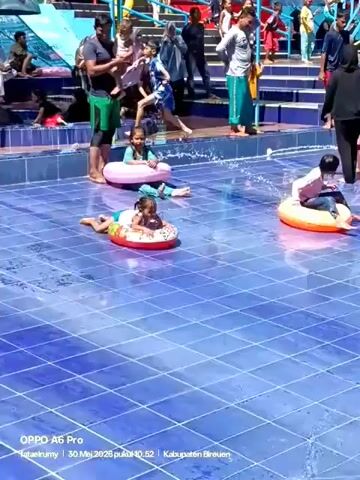The video shows a group of people, including children and adults, enjoying themselves at a water park. The scene focuses on a shallow, blue-tiled pool where several children are playing with inflatable tubes. They are lying on their stomachs, using their hands to paddle and move around the pool. In the background, there are more people walking, standing, and interacting, some near a small slide and others around the pool's edge. One adult woman wearing a hijab and dark clothing is sitting at the edge of the pool, assisting a child in an inflatable tube. The overall atmosphere is lively and cheerful, with people enjoying a sunny day at the water park.  A time stamp and phone model appear at the bottom of the video.