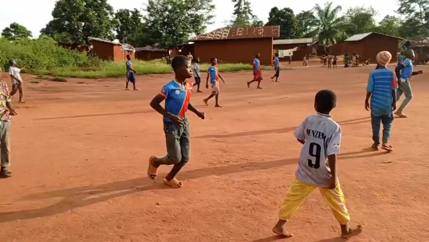 The video shows a group of children playing soccer on a dirt field in what appears to be a rural village. The field is surrounded by trees and small, simple houses made of reddish-brown material. Some of the children are wearing soccer jerseys, while others are in casual clothing. The children are running and kicking a soccer ball, seemingly engaged in a game. The camera pans across the field, capturing the various players and their activities. The overall atmosphere is one of youthful energy and playful activity.
