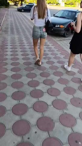 Two young women walk away from the camera on a patterned sidewalk. The woman on the left wears a white top and denim shorts, carrying a black purse. The woman on the right wears a black dress. They walk on a sidewalk with a pattern of red circles and gray squares. Cars and trees are visible in the background. The scene appears to be outdoors during the daytime.