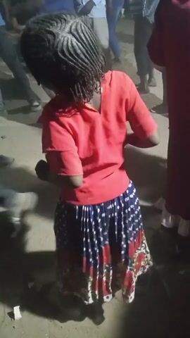The video shows a young girl with braids, wearing a red t-shirt and a colorful skirt, dancing in an outdoor setting. Several other people are visible in the background, some standing and watching. The ground appears to be dirt or sand. The lighting is dim, possibly dusk or evening. The girl is the primary focus, with her movements suggesting she is enjoying the music.