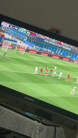 The video shows a television screen displaying a soccer match. The game is between France (FRA) and Morocco (MAR). The score is 0-0, and the time is 27:48. A player in a white uniform, identified as Mbappé, is taking a shot at the goal. The goalkeeper, wearing a lime green jersey, dives to make a save, and successfully blocks the shot. The crowd in the stadium is visible in the background. The FIFA World Cup 2026 logo is also displayed on the screen. The television is a Samsung brand. The scene is viewed from a slight angle, with a patterned tablecloth visible in the foreground.
