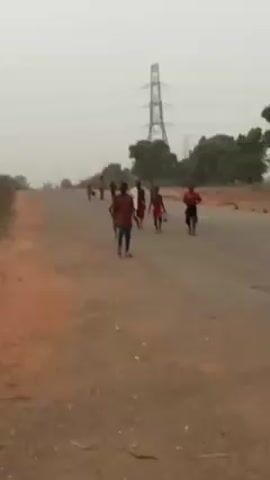 A medium shot shows a group of at least nine children walking or running on a dirt road. They are of various sizes, and several are shirtless. The child closest to the camera wears a red, patterned shirt with blue pants. Some carry what appears to be a small container or plastic bag. An electrical tower is visible in the background, along with sparse trees and an overcast sky. The overall impression is one of a rural or less developed area.
