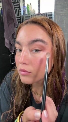 A fair-skinned woman with light brown hair is applying makeup to her left eyelid with an eyeshadow brush. She is facing the camera and looking into a mirror that is visible in the background. Her hair has hints of orange and streaks of purple tinsel. She is wearing a black t-shirt and gold bangles on her left wrist. The bathroom behind her features a black and white patterned wall, a towel hanging on a rod, and various toiletries on a shelf.