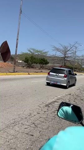The video presents a first-person view from a motorcycle traveling along a paved road under a clear blue sky. The road appears to have two lanes, one for each direction, separated by a yellow curb. The road surface directly in front of the motorcycle shows noticeable cracks.

In the foreground, parts of the motorcycle are visible, including the rearview mirror on the right, which reflects a person wearing a teal shirt. Text like "Ride" and "sidis" can be seen on the mirror or a helmet. A phone is mounted on the handlebars, showing what appears to be a map or navigation interface.

Ahead, a silver hatchback car is driving in the same direction. Its license plate is partially visible, showing "1449339". Further down the road and to the left, another motorcycle is visible, moving around a gentle curve.

The surrounding landscape features a mix of vegetation. On the left side of the road, there's a grassy strip with several green trees and some dry, bare trees with widespread branches. In the background, hills or mountains with sparse vegetation are visible under the bright sunlight. Power lines stretch across the sky. The overall setting appears to be a sunny day in an area consistent with Los Cerritos, Venezuela.