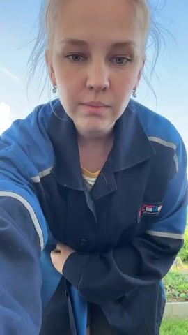The video begins with a close-up of a young woman's face. She has blonde hair tied up in a bun and is wearing a blue and navy work jacket. She then turns and stands up, the camera panning out to reveal her standing in what appears to be a garden. She smiles and adjusts her jacket. Another person, a man, enters the frame from the right and stands behind the woman. He puts his arms around her and they both smile at the camera. The environment appears to be outdoors on a sunny day with a clear blue sky and some clouds. There are garden beds with plants and trees in the background, as well as a building and a fence.