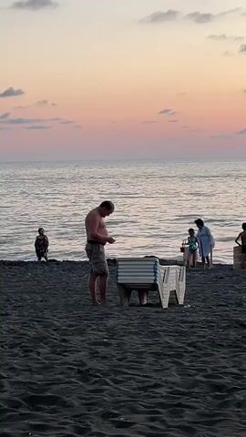 The video shows a beach with black sand and calm ocean waves. The sky is a soft gradient of pink and orange, indicating either sunrise or sunset. In the foreground, a man wearing shorts is bending over a stack of white lounge chairs. Further back, several other people are visible near the water. To the left, a woman stands facing away from the camera. In the center, a woman in a white garment and a child are visible, and to their right, another child stands. The overall atmosphere is peaceful and serene.