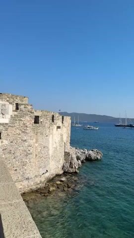 The video pans from left to right across a body of water. In the foreground on the left, there is a stone wall and part of a building with square openings. The wall is constructed of rough-hewn stones. To the right of the wall is the clear, blue-green water, with rocks visible beneath the surface near the shore. Several boats are anchored or sailing in the water, including sailboats and smaller motorboats. In the background, a hilly landmass with vegetation is visible across the water. The sky is clear and blue.