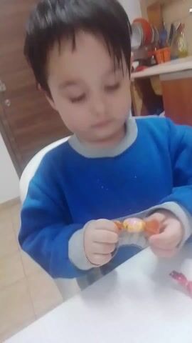 The video shows two children in what appears to be a kitchen. The first child, a young boy with dark hair wearing a blue sweatshirt, is seated at a white table and is in the process of unwrapping a piece of candy.  The second child, a young girl with dark hair wearing a light pink Paw Patrol patterned sweater, is standing and also unwrapping a piece of candy. The kitchen has tan tile flooring.  In the background, there is a doorway, white sneakers, a countertop with kitchen utensils, and a bottle of dish soap.