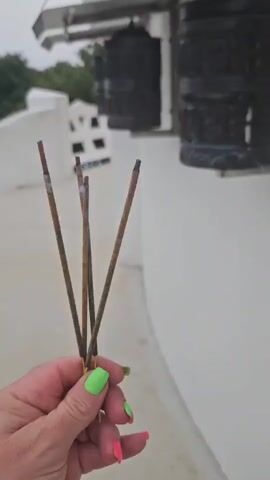 A hand with brightly painted nails holds three incense sticks that are burning and producing smoke. In the background, there are prayer wheels mounted on a white structure, with trees visible beyond.
