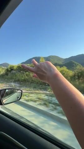 This video captures a first-person perspective from inside a moving vehicle, looking out the passenger window. A person's arm and hand are visible, extending out of the open window. The background shows a scenic landscape with rolling green hills and trees under a clear blue sky. A road with a white line and a guardrail is visible alongside the car, along with a side-view mirror. The person's hand makes a series of gestures, including clenching into a fist, opening into a palm, and forming a hand gesture that resembles the "rock on" sign. The overall impression is one of enjoying a scenic drive.