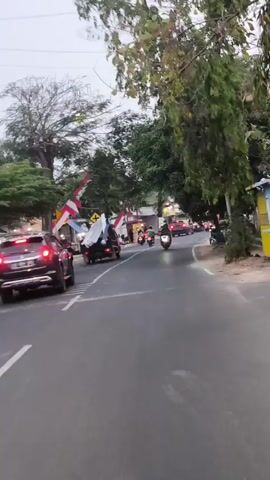 The video shows a street scene with several vehicles and people. There are trees lining the sides of the road, decorated with red and white flags. A dark-colored SUV is in the middle of the road, and there are several motorcycles and cars moving in the same direction. Some people are riding on the motorcycles. On the sides of the road, there are buildings that appear to be shops or businesses. The sky is overcast, suggesting it might be dawn or dusk. The overall impression is of a busy street, possibly during a local event or celebration, given the flags.