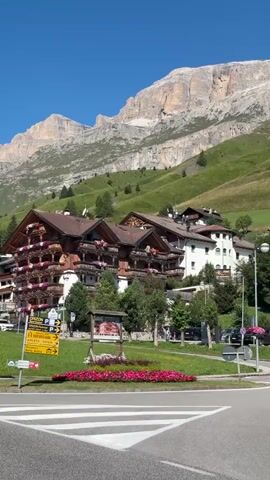 The video shows a village nestled in a valley with a large, rocky mountain in the background. The village features traditional wooden chalets with balconies adorned with flowers. A road curves through the scene, with cars parked along the side. The sky is clear and blue, and the sun is shining, casting shadows on the grassy slopes of the mountain. The overall atmosphere is peaceful and scenic.