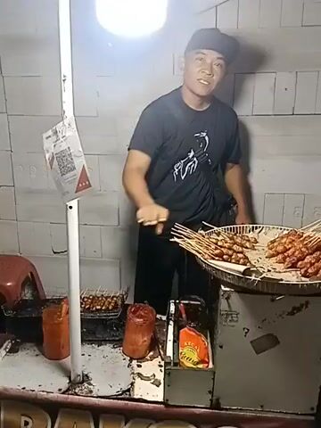 The video shows a man standing behind a food cart. He is wearing a black t-shirt with a graphic design and a black cap. He is holding a skewer of food. In front of him on the cart, there are more skewers of food, a grill, bottles of sauce, and a QR code displayed on a stand. The background appears to be a wall made of cinder blocks. The lighting is bright, coming from a circular light fixture above the man's head. The man is looking towards the camera.