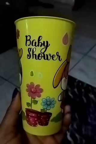 A hand holding a yellow plastic cup with the words "Baby Shower" written on it. The cup has a drawing of a cartoon dog character wearing a bow and glasses, and a flower pot with two flowers. The background is a tiled floor with a black-and-white rug.