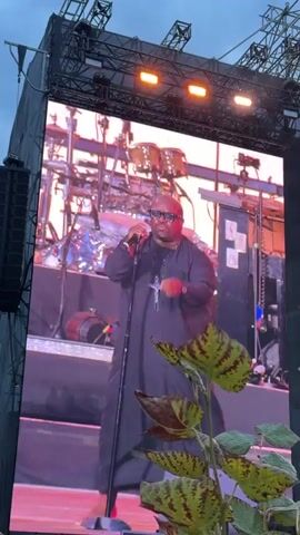 The video shows a concert scene. A large screen displays a drummer playing, then switches to a smiley face logo. The stage has lights and several performers. The crowd is visible in the foreground. The overall environment appears to be outdoors with a cloudy sky.