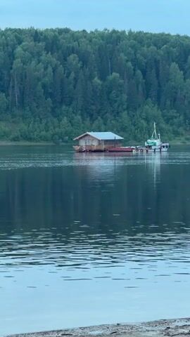 A floating house with a boat attached to it is on a large body of water. The water is calm and reflects the trees on the shore. The trees are dense and green, and they form a backdrop to the scene. The sky is overcast and gray. The water is the foreground of the image, and it is rippled. The floating house is in the middle ground, and it is a simple wooden structure with a metal roof. The boat is a small white vessel with a green cabin. The trees are in the background, and they are lush and green. The sky is a muted gray, and it is overcast.
