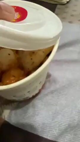 The video shows a paper cup filled with a food dish. The food appears to be a mixture of fried dough pieces, small white spherical items, and a red-tinged broth. The paper cup is placed on a surface that looks like a bed or a cushioned surface with a gray and white pattern. In the background, out of focus, there's a dark-colored pouch or case and what appears to be a white electronic device. The video begins with a white lid being lifted to reveal the dish.