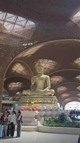 This is a video of the interior of a large, modern airport terminal. The architecture is notable for its sweeping, arched roof made of woven wood or bamboo, with skylights allowing natural light to filter through. In the foreground, a large, golden Buddha statue sits on a raised platform adorned with flowers. Several people, including a woman holding a child, are visible near the statue. As the camera pans and moves, the video shows the bustling airport environment. Travelers with luggage are seen walking through the terminal. Check-in counters, information boards, and shops are discernible in the background. The overall atmosphere is one of movement and activity typical of an airport.