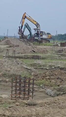 This is a vertical, eye-level, medium shot of a construction site. In the background, two excavators are working. The excavator on the left is yellow and the excavator on the right is green and white. Both excavators are digging up dirt. In the foreground, there is a metal cage made of rebar, which is likely used for reinforcing concrete. The ground is muddy and uneven, with piles of dirt and some grass. The sky is overcast. The scene suggests that construction is in progress.