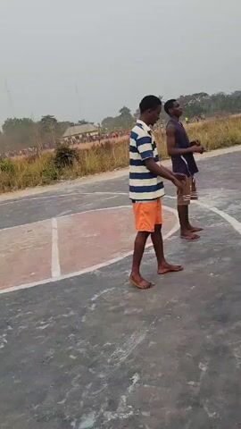 Two young men are on a basketball court in an outdoor, sunny setting. One is wearing a striped blue and white shirt with orange shorts, while the other is in a dark blue tank top and dark brown shorts. The man in the striped shirt performs a backflip while the other man steps back slightly. After landing, the man in the striped shirt smiles and walks towards the camera. The basketball court has a gray surface with white lines and a painted red circle in the center. In the background, there are trees and a crowd of people gathered on a field.