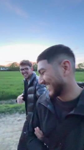 Two men are walking on a dirt path that runs next to a large field of green grass. The man in front is wearing a black hooded sweatshirt and dark pants. The man behind him is wearing glasses, a black puffer jacket, and dark pants. The sky is a mix of blue and pink, suggesting it is either early morning or late evening. They are walking closely together, and at one point the man behind puts his arm around the other's shoulders. The focus and clarity are affected by movement.