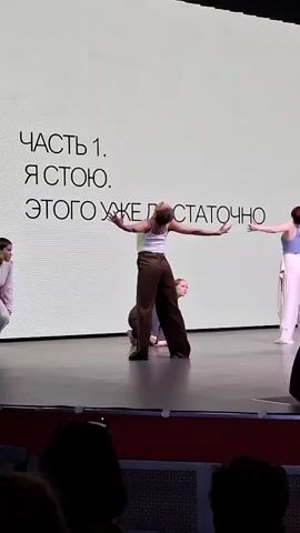 The video shows a dance performance. A large screen at the back of the stage displays the text "Часть 1. Я стою. Этого уже достаточно." There are five dancers on the stage. One dancer is in a white top and brown pants, another in a light blue top and white pants. The other three dancers are wearing less visible clothing, but one is wearing a dark colored top. All five dancers are moving in various poses, and two of them are on the ground. There is a small audience visible in the foreground. The stage is dark, and there is a red trim along the front of the stage.