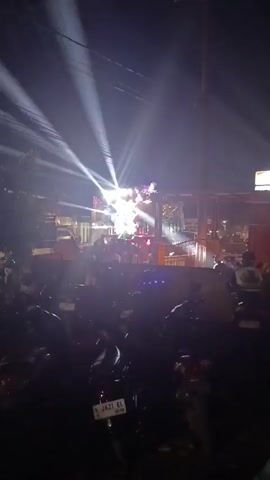 The video shows a night scene with a crowd gathered in front of a stage illuminated by bright spotlights. Numerous motorcycles are parked in the foreground, partially obscuring the view of the crowd. The stage appears to be set up outdoors, possibly on a street or in an open area, with buildings and structures visible in the background. The overall atmosphere suggests an event or performance taking place.