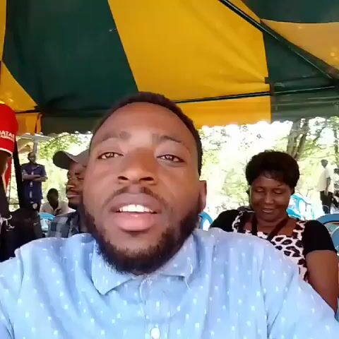 Here is a brief description of the video:

The video shows a man sitting under a canopy, possibly at an outdoor event. The canopy is yellow and green.  A woman sits to his right, looking at her phone. Other people are visible in the background, seated in blue plastic chairs. The background appears to be outdoors, with trees visible. The man in the foreground is looking into the camera and appears to be talking, likely recording himself. There is music playing in the background.