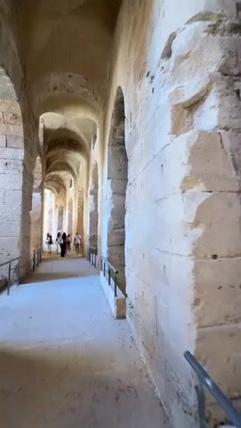 The camera moves slowly down a long, vaulted hallway of an ancient stone building. The hallway is illuminated by light from the far end, and it has a smooth concrete floor. Arches line both sides of the hallway, supported by thick stone pillars. A metal railing runs along the edge of the hallway. Several people are walking towards the end of the hallway, including women in dresses. The stone walls show signs of age and wear. The sky is visible through the arches.