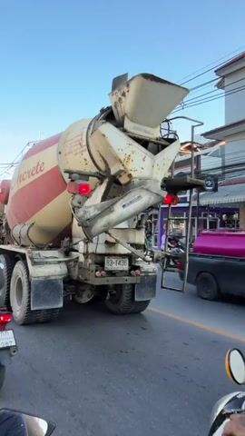 The video shows a concrete mixer truck driving on a road. The truck is large and white with red stripes and the word "Concrete" written on the side. The truck is moving alongside other vehicles, including cars, vans, and motorcycles. The sky is blue and clear, and there are power lines and trees visible in the background. The video appears to be filmed from another vehicle, possibly a motorcycle, as indicated by the handlebars and a portion of the rider's arm in the foreground. The scene appears to be in a city or town, with buildings and signs visible along the roadside. The traffic seems to be moderate, with vehicles moving at a slow pace.