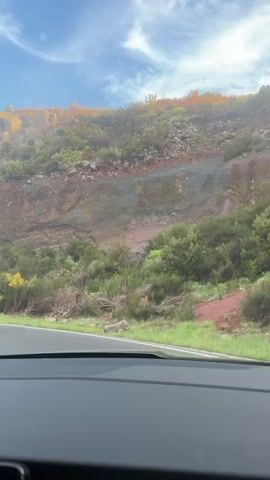 A vehicle travels on a winding road. The view is from inside the car, looking through the windshield. The road is paved and has white lines marking the lanes. There are yellow poles on the roadside. On the right side of the road is a hill covered with vegetation and rock formations. The sky above is blue with scattered clouds. The dashboard of the car is visible at the bottom of the frame. The car is moving uphill, as indicated by the road's incline. The overall impression is that the vehicle is traveling through a mountainous area on a sunny day.