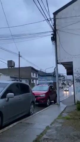 The video shows a street scene on a wet, overcast day. Several cars are parked along the sidewalk on the left side of the frame, including a silver hatchback and a red SUV.  Other cars are visible further down the street, driving on the wet road. Buildings line the street, including a two-story grey house with white-trimmed windows and a white building to the right, which has exposed graffiti near the bottom. A utility pole stands on the right side of the frame, surrounded by a tangle of overhead wires. The sidewalk and street are wet, reflecting light and indicating recent or ongoing rain. The sky is grey and overcast, suggesting a gloomy atmosphere.