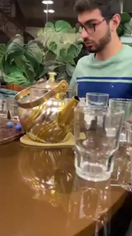 Three young men sit around a shiny, wooden table in what appears to be a restaurant or cafe. The focus shifts between them, showing each at different moments. The first man wears glasses, a beard, and a green shirt. He is near a silver and gold teapot and several clear glasses. The second man, wearing a gray shirt, holds playing cards and appears surprised. A wooden game board with marbles is in front of him. The third man, in a white shirt, appears bored or tired. Empty glasses and a cigarette packet sit near him. The environment includes lush green plants in the background. The lighting is warm and bright.