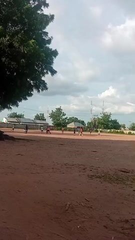 A group of children are playing soccer on a dirt field. A large, red and white telecommunications tower stands tall in the background. The sky is cloudy, and trees are visible in the distance.